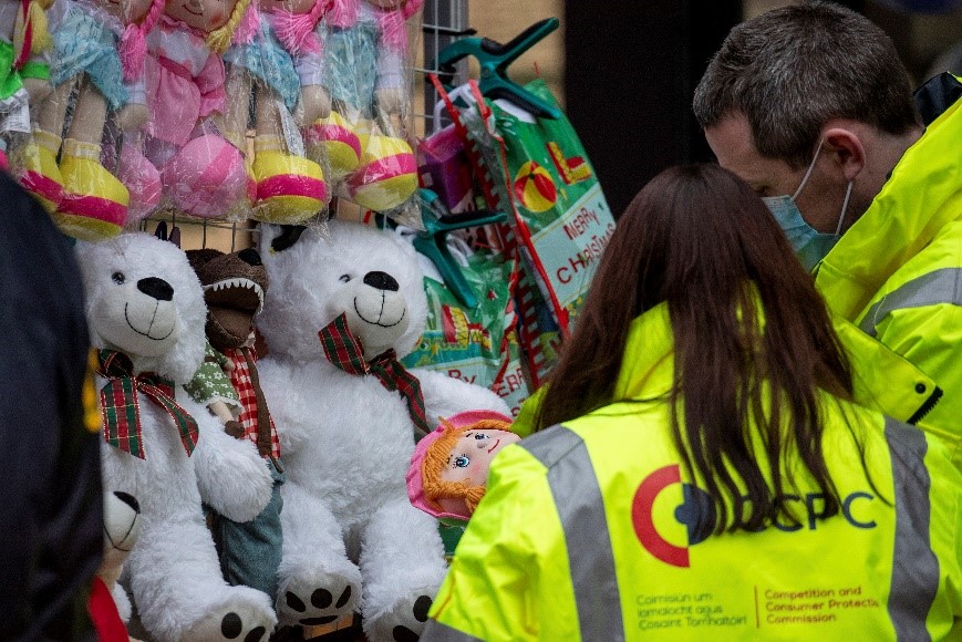 Two CCPC Authorised Officers in branded hi-vis jackets inspecting children’s toys for sale at an outdoor market stall.