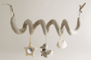 Soft, fabric spiral pendant designed for stroller attachment, made of light beige and white checkered material. It features three hanging toys: a star with a mirror center, a duck, and a cloud. The spiral wraps around the stroller handle or bar, secured with ties at both ends.