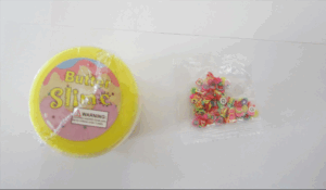 Yellow and round container with Butter Slime written on top. Small plastic bag containing small plastic pieces on the right.