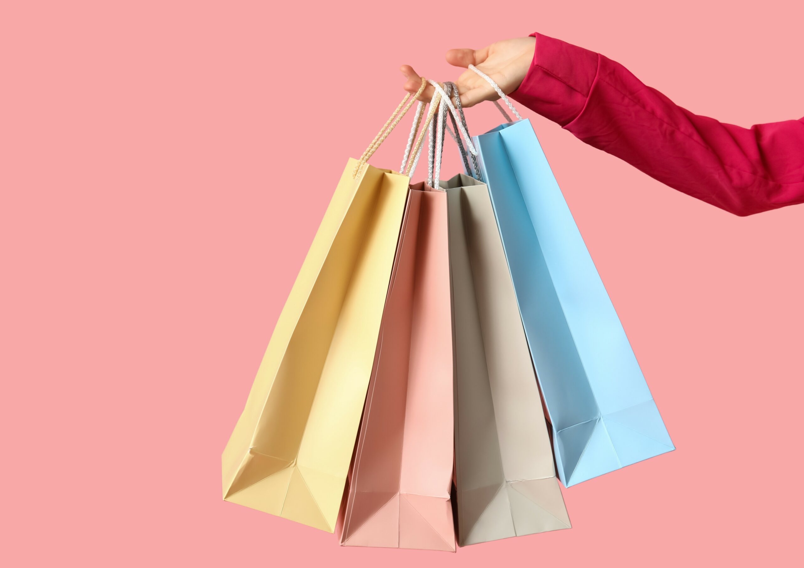 A hand holding out four pastel-coloured shopping bags against a pink background.