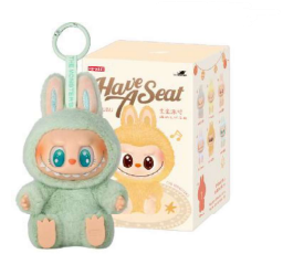 A plush toy figure dressed in a mint-green bunny costume with large blue eyes and a wide smile, holding a circular handle on top. Behind the toy is a rectangular box featuring an illustration of a similar character in a yellow bunny outfit and the text ‘Have A Seat’ along with smaller decorative elements and product details.