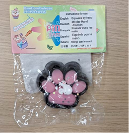 A packaged squishy toy shaped like a pink paw with black edges and white pads, sealed in clear plastic. The top of the package has a colorful label with text in multiple languages. The English instructions read: “Squeeze by hand.” Other languages include German, French, Spanish, and Italian. The label also has playful graphics and phrases like “Emotional release” and “Relieve stress.”