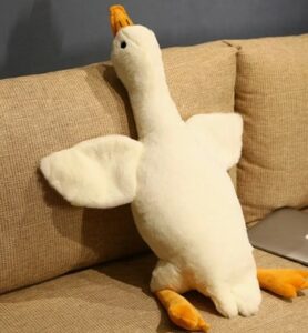A plush toy shaped like a white duck with orange beak and feet, sitting upright on a beige fabric couch. Its wings are spread outward, and part of a silver laptop is visible on the couch beside it.