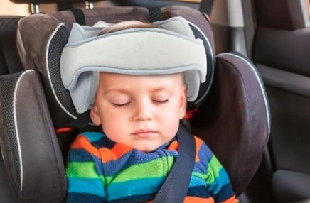 A young child asleep in a car seat with a grey head strap on its head.