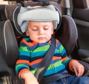 A young child asleep in a car seat with a grey head strap on its head.