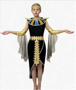 A child standing with arms extended outward while wearing an Egyptian‑inspired costume. The outfit includes a black knee‑length garment with gold trim, a wide decorative collar featuring blue and gold accents, gold arm bands, and sheer black draped fabric attached at the arms. A gold belt with geometric detailing hangs down the front