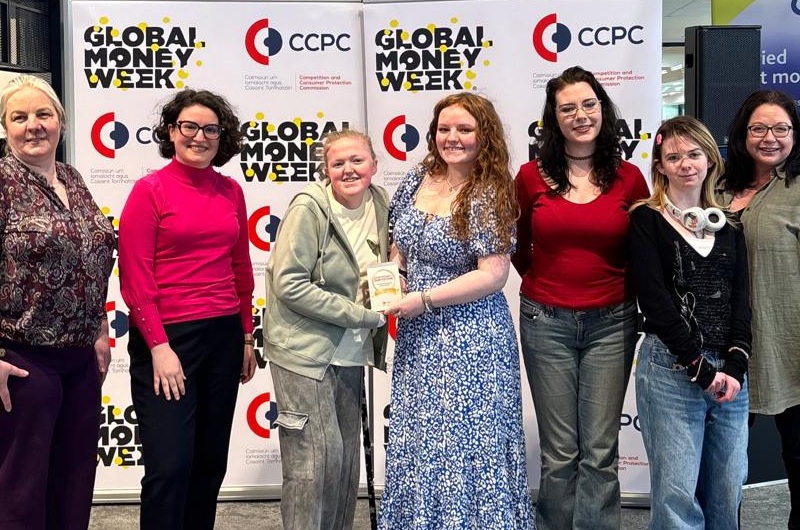 A group of students and educators stand in front of Global Money Week banners holding an award.
