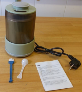 A compact electric grain mill in light green and transparent plastic. A black power cord is attached to the mill. In front of the mill are a small blue cleaning brush, a white plastic measuring spoon, and a folded instruction leaflet.
